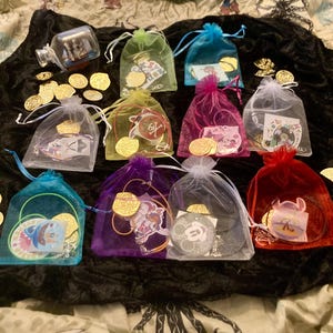 May include: Ten small, colorful, sheer bags filled with gold coins and charms. Each bag has a different design and color, including blue, purple, red, and green. The bags are arranged on a black fabric surface.