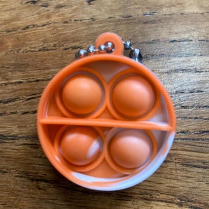May include: A white and orange pop it fidget toy with a silver chain attached. The toy has four large bubbles in a circular shape.