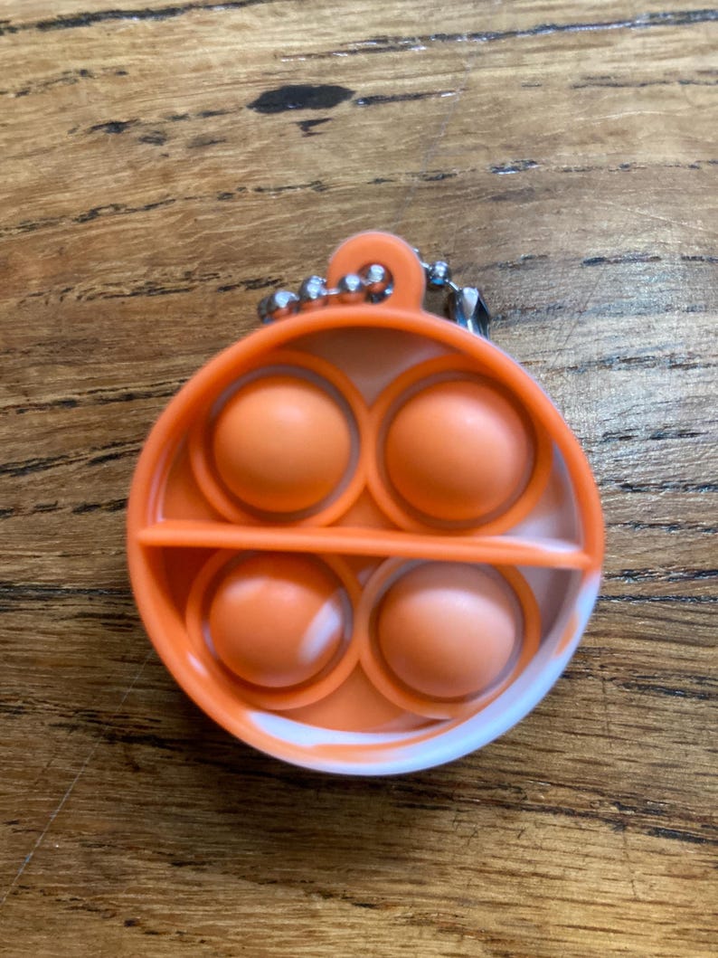 May include: A white and orange pop it fidget toy with a silver chain attached. The toy has four large bubbles in a circular shape.