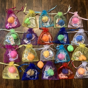 May include: Twenty-four small, colorful, rubber duck toys are displayed in individual, sheer, organza bags. Each bag is tied with a ribbon and contains a different colored duck. The bags are arranged in four rows of six bags each.