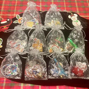 May include: A collection of Christmas ornaments in clear, snowflake-patterned bags. The ornaments feature various Disney characters and holiday-themed designs. The bags are tied with silver ribbons and arranged on a black surface, with additional decorative ornaments around.