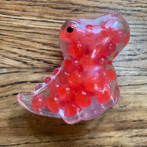 May include: A clear plastic dinosaur toy filled with red squishy balls. The dinosaur has a black eye and a pink mouth.