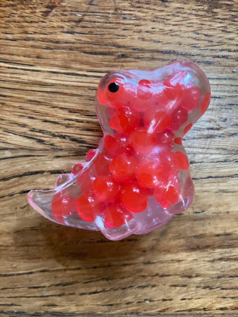 May include: A clear plastic dinosaur toy filled with red squishy balls. The dinosaur has a black eye and a pink mouth.