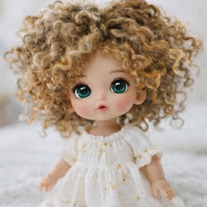 Puede incluir: Una muñeca con grandes y expresivos ojos verdes y mejillas rosadas. La muñeca tiene cabello rubio rizado y viste un vestido blanco con detalles florales amarillos. El vestido tiene mangas cortas y un dobladillo con volantes. Es un objeto de colección encantador.