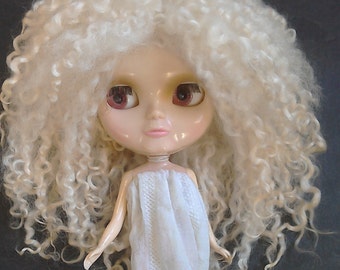 Op de afbeelding: Een pop met een bleke teint, grote ogen en een serene uitdrukking. De pop heeft lang, krullend, lichtblond haar en draagt een wit kledingstuk. De ogen van de pop zijn roodbruin van kleur. De pop is een verzamelobject.