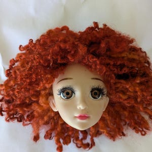 May include: A doll head with a fair complexion, brown eyes, and long eyelashes. The doll has a small, pink mouth and a full head of vibrant, curly red hair. The doll's face is centered, and the background is a plain white surface.
