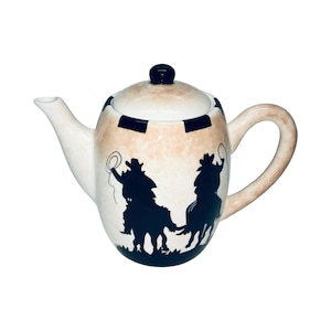 May include: Cream-colored ceramic teapot with a lid and handle. The teapot features a tan and white speckled design with black silhouettes of cowboys on horseback. The lid has a black knob and black stripes.