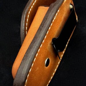 May include: A brown leather belt holster with a black metal clip. The holster is designed to hold a small object, such as a knife or a flashlight.