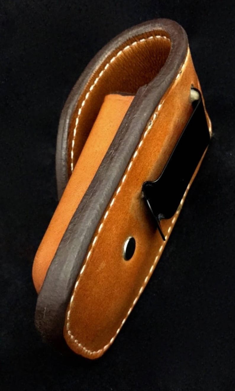 May include: A brown leather belt holster with a black metal clip. The holster is designed to hold a small object, such as a knife or a flashlight.