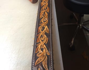 May include: A brown leather guitar strap with a detailed floral design carved into the leather. The design is in a light brown color and features a small wolf image at the bottom.