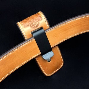 May include: A brown leather belt with a stitched edge and a black metal clip. The belt is attached to a brown leather holster with a stitched edge and the word "NAY" embossed on it.