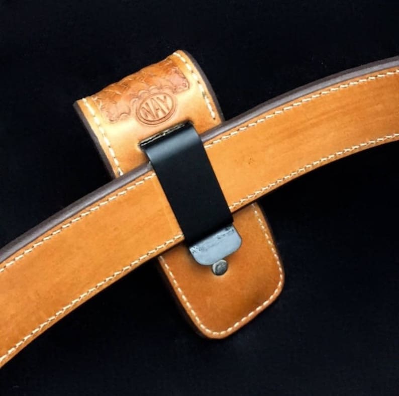 May include: A brown leather belt with a stitched edge and a black metal clip. The belt is attached to a brown leather holster with a stitched edge and the word "NAY" embossed on it.