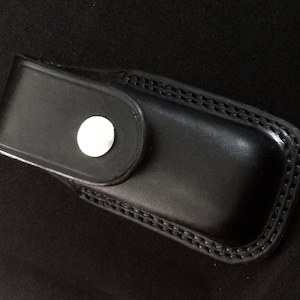 May include: A black leather case with a snap closure and a white button. The case is designed to hold a small pocket knife or other similar tool.