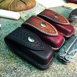 May include: Three leather knife sheaths with a decorative stitched pattern. The sheaths are black, burgundy, and brown. A silver multi-tool is also pictured.