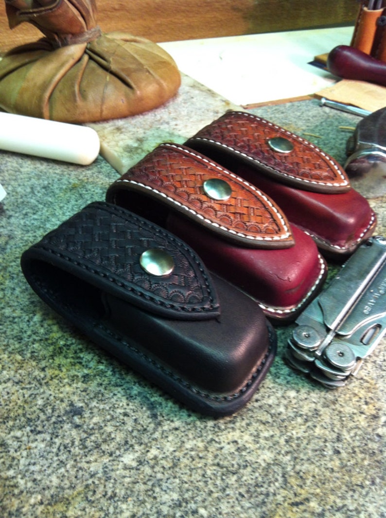 May include: Three leather knife sheaths with a decorative stitched pattern. The sheaths are black, burgundy, and brown. A silver multi-tool is also pictured.