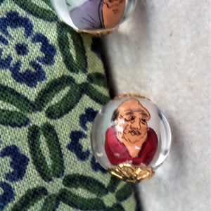 May include: Two glass beads with gold trim, each containing a painted portrait of a person with a white background.