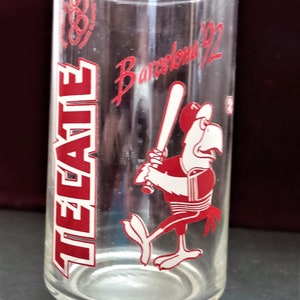 May include: A clear glass with a red and white design. The glass features the text "Tecate" and "Barcelona '92". A cartoon rooster holding a baseball bat is also featured on the glass.