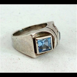 May include: A silver ring featuring a square, light blue gemstone in a square setting. The ring has a thick band and additional dark blue stones on the side.