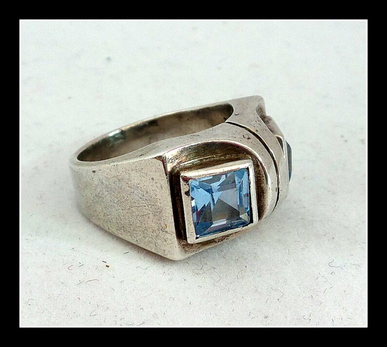 May include: A silver ring featuring a square, light blue gemstone in a square setting. The ring has a thick band and additional dark blue stones on the side.