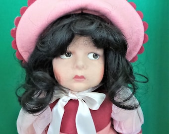 May include: A vintage doll with black hair, wearing a pink hat with a red scalloped brim and a pink rose. The doll has a pink dress with a white ribbon and a red bodice. The doll's face is painted with rosy cheeks and lips.