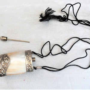 May include: A white and silver ornate bottle with a black cord and tassel. The bottle has a silver cap and a small silver tube.