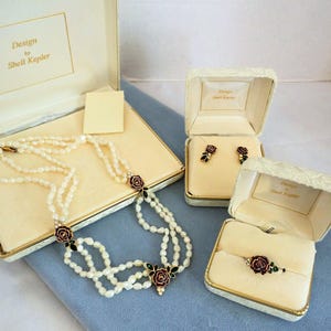 May include: A vintage jewelry set by Shell Kepler, featuring a multi-strand pearl necklace, earrings, and a bracelet. The set includes rose-shaped accents with red and green enamel details, presented in cream-colored boxes.