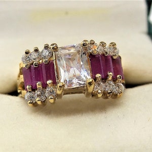 May include: A gold ring with a large, clear, square-shaped center stone surrounded by smaller clear stones and red baguette-shaped stones.