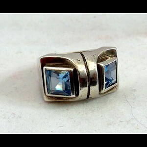 May include: A silver ring featuring two square, light blue gemstones. The ring has a geometric design with a rectangular band and square settings for the gemstones. The gemstones are a vibrant, clear blue colour.