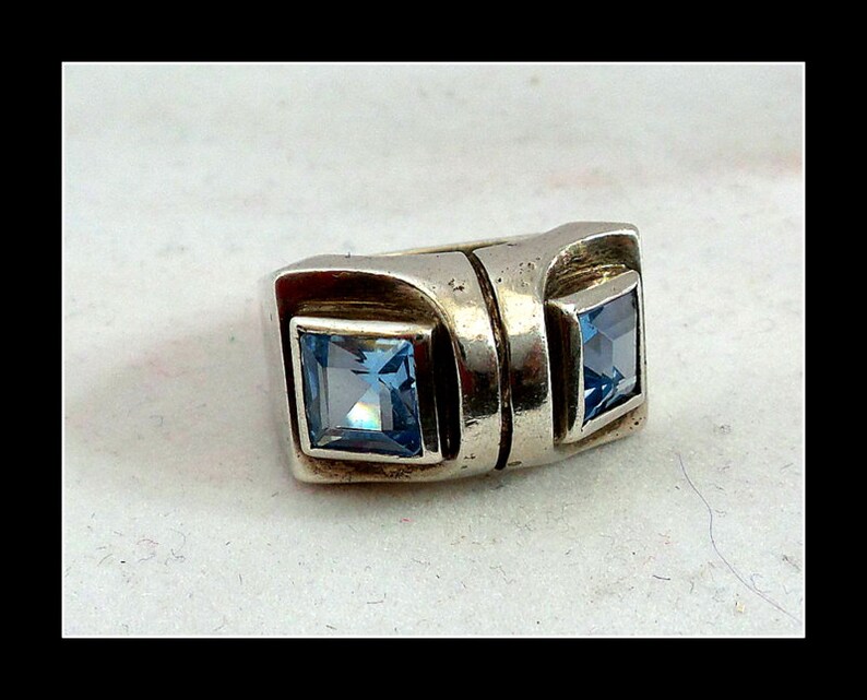 May include: A silver ring featuring two square, light blue gemstones. The ring has a geometric design with a rectangular band and square settings for the gemstones. The gemstones are a vibrant, clear blue colour.