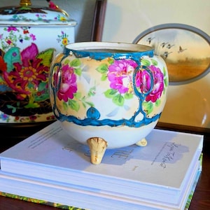 May include: A round, three-footed porcelain planter featuring a cream-coloured base, vibrant pink floral patterns, and a decorative teal blue border with gold accents. The planter has a wide opening and sits atop a stack of two hardcover books.