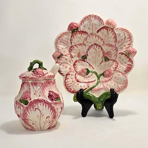 Peut inclure: Un pot en céramique avec couvercle et une assiette décorative présentant un motif en relief de fraises et de feuilles. Les deux pièces sont vernies en crème avec des détails peints à la main en rose et rouge, posées sur un support noir.