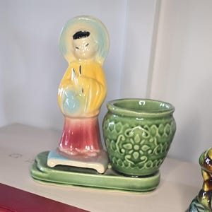 Puede incluir: Juego de figuras de cerámica vintage. Presenta una figura con una túnica amarilla con un halo azul y una maceta verde. La figura está sobre una base verde con una falda roja y rosa. La maceta tiene un diseño floral.