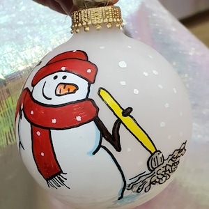 May include: White glass Christmas ornament with a hand-painted snowman wearing a red scarf and hat, holding a yellow broom. The background is white with white dots.