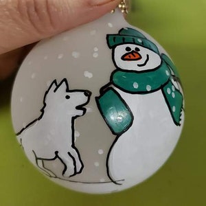 May include: A hand-painted Christmas ornament featuring a snowman and a dog. The snowman wears a green hat and scarf, and the dog is white with black outlines. The ornament has a light gray background with white snowflakes.