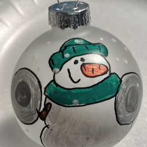 May include: A white glass Christmas ornament with a hand-painted snowman wearing a green hat and scarf. The snowman is holding two gray dumbbells.