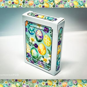 Könnte beinhalten: Ein Kartenspiel mit hellblauem Hintergrund und einem Design aus bunten Ostereiern und Blumen. Die Schachtel trägt die Aufschrift "Easter Flowers Playing Cards".