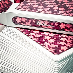May include: A deck of playing cards with a pink and white floral design. The box features the word "Sakura" in cursive.
