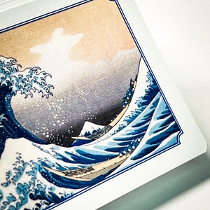 May include: A close-up of a playing card featuring a blue and white image of a large wave crashing against a mountain. The card has a blue and white border.