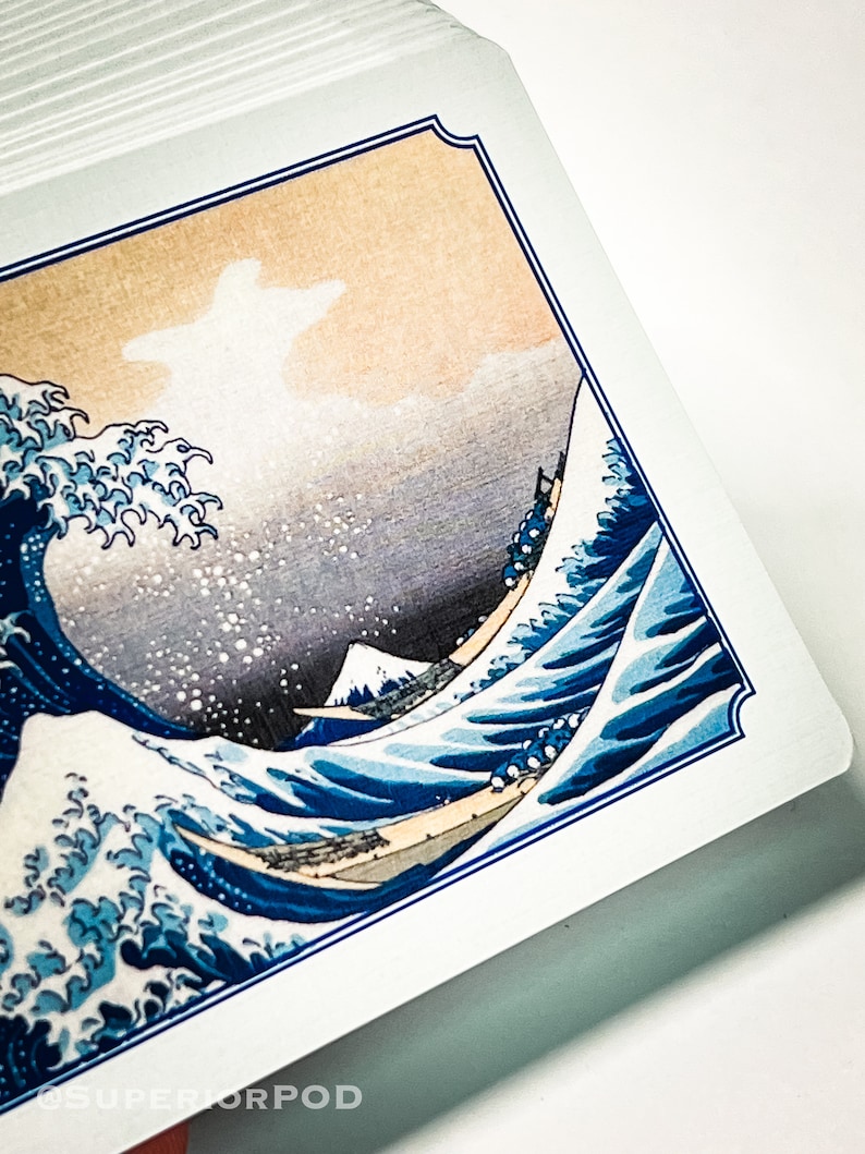May include: A close-up of a playing card featuring a blue and white image of a large wave crashing against a mountain. The card has a blue and white border.