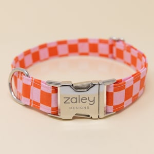 Op de afbeelding: Een roze en oranje geruite hondenriem met een zilveren gesp waarop "zaley designs" staat.