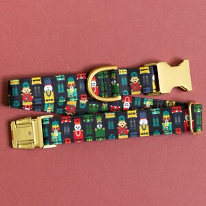 May include: A black dog collar with a gold buckle and a festive pattern of nutcracker soldiers. The collar is made of fabric and has a repeating pattern of nutcracker soldiers in various colors, including red, green, blue, and yellow.