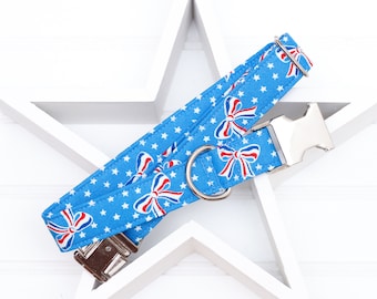 Puede incluir: Un collar de perro azul con estrellas blancas y lazos rojo, blanco y azul. El collar tiene una hebilla plateada y un anillo en D plateado.
