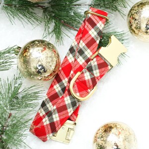 Op de afbeelding: Rode en witte geruite hondenriem met gouden beslag. De riem ligt op een wit oppervlak met gouden ornamenten.