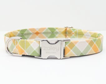 May include: A dog collar with a plaid pattern in shades of yellow, green, and white. The collar has a silver-colored metal buckle and a D-ring. The brand name "Zaley Designs" is printed on the buckle.