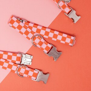Op de afbeelding: Drie hondenhalsbanden met roze en oranje ruitjespatroon en zilveren gespen. De halsbanden hebben de tekst "Zaley Designs" op een van de gespen.