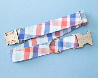 May include: A blue, pink, and white plaid dog collar with a silver buckle and a silver ring. The buckle has the text "zaley designs" engraved on it.