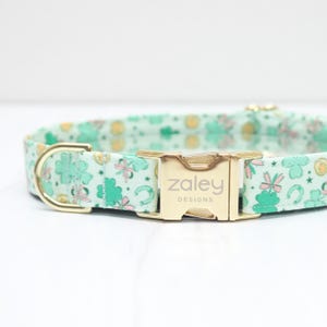 May include: A light green dog collar with a gold buckle. The collar has a pattern of shamrocks, horseshoes, and stars. The buckle has the text "zaley designs" engraved on it.