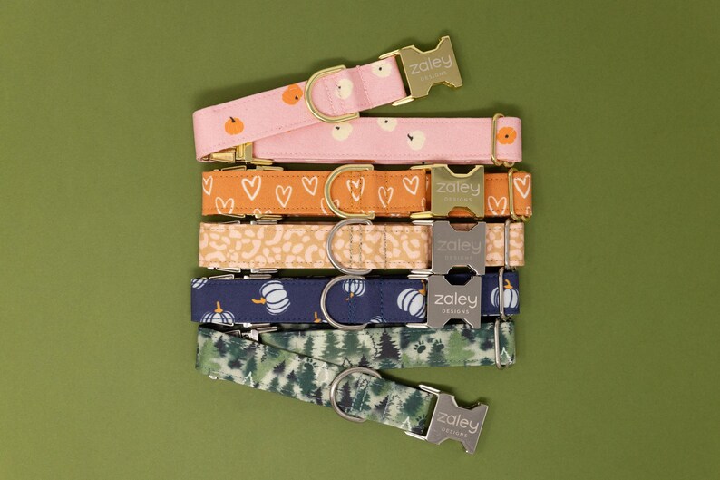 May include: A stack of six colorful dog collars with different patterns, including pumpkins, hearts, and leaves. Each collar has a silver buckle with the text "Zaley Designs".