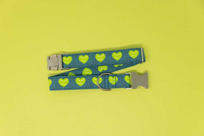 May include: A teal dog collar with a pattern of lime green tennis ball hearts. The collar has a silver metal buckle and a D-ring. The background is a solid yellow-green.
