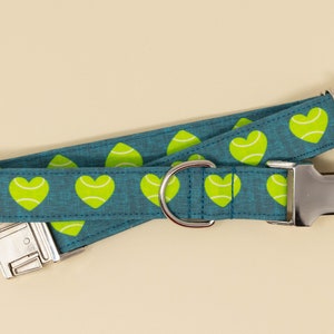 May include: A teal dog collar with a repeating pattern of lime green hearts. The collar has a silver metal buckle and a D-ring for leash attachment. The background is a solid beige color.
